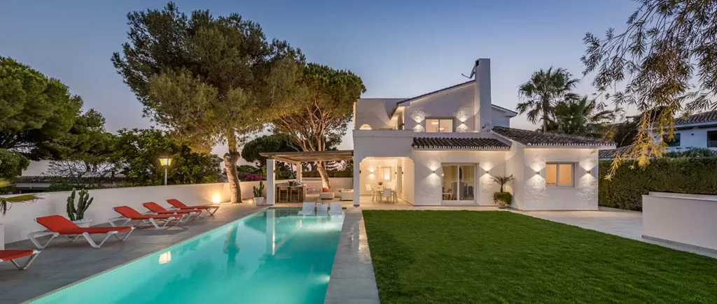 Modern white villa with a pool, surrounded by greenery and lounge chairs.
