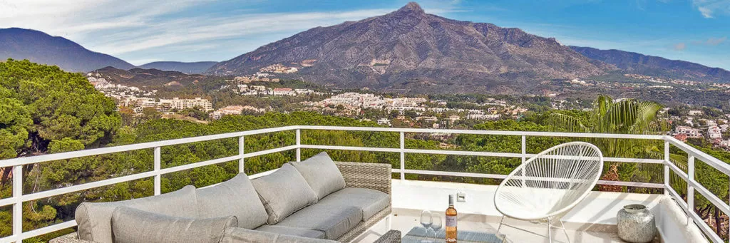 A modern terrace overlooks a mountainous landscape, featuring comfortable seating and a drink.